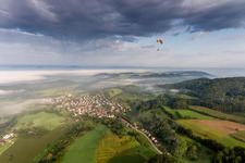 Paragleiter über Dorf - Ansicht am Rande von Feldern vor dem von Frühnebel verhüllten Bodensee im Ortsteil Güttingen in Radolfzell am Bodensee im Bundesland Baden-Württemberg, Deutschland